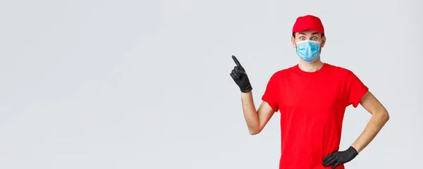 Covid-19, self-quarantine, online shopping and shipping concept. Surprised delivery guy popping eyes amazed or excited as showing promo, pointing finger left, wear courier uniform and medical mask — Stock Photo, Image