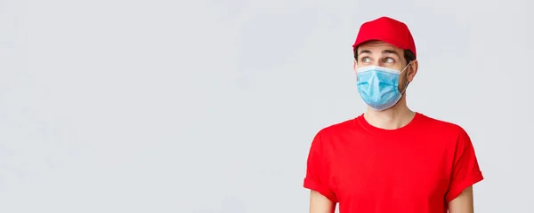 Contactless delivering, covid-19 and shopping concept. Thoughtful courier in red uniform cap, t-shirt and medical face mask with gloves, looking upper left corner, reading banner or advertisement — Stock Photo, Image