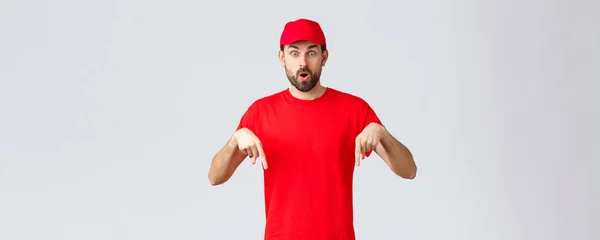 Compras en línea, entrega durante la cuarentena y concepto de comida para llevar. Sorprendido e intrigado mensajero informar a los clientes, empleado en gorra uniforme rojo y camiseta señalando los dedos hacia abajo, decir wow —  Fotos de Stock