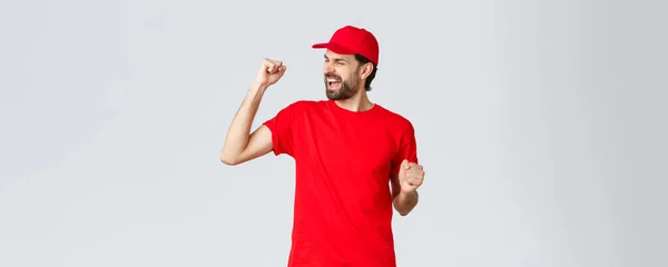 Fröhlicher und unbeschwerter junger bärtiger Lieferant in roter Uniformmütze und T-Shirt, singend, johlend und faustdick in Feierlaune, triumphierend und jubelnd über großartige Neuigkeiten, grauer Hintergrund — Stockfoto