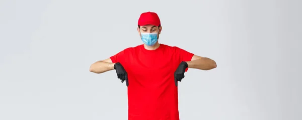 Covid-19, self-quarantena, shopping online e concetto di spedizione. Curioso corriere che punta e guarda in basso, indossando maschera e guanti medici, protegge i dipendenti durante l'epidemia di coronavirus — Foto Stock