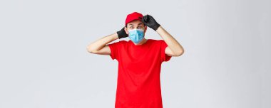 Market ve paket teslimatı, covid-19, alışveriş konsepti. Heyecanlı kırmızı üniformalı teslimatçı, kafasına şapka takıyor ve ilgilenen kameraya bakıyor, koronavirüs sırasında kurye görevinde.