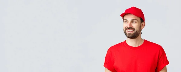 Achats en ligne, livraison en quarantaine et concept à emporter. gai barbu guy en rouge uniforme cap et t-shirt, regarder loin avec sourire heureux, lecture bannière signe, fond gris — Photo