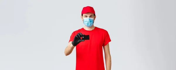 Contactless delivery, payment and online shopping during covid-19, self-quarantine. Handsome courier in red uniform, cap, medical face mask and gloves, show credit card, order internet — Stock Photo, Image