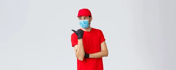Covid-19, αυτο-καραντίνα, online αγορές και ναυτιλία έννοια. Ο τύπος παράδοσης με την κόκκινη στολή, γάντια και μάσκα προσώπου, δείχνοντας αριστερά και χαμογελώντας, συστήνει την υπηρεσία κούριερ χρήσης πελάτη — Φωτογραφία Αρχείου