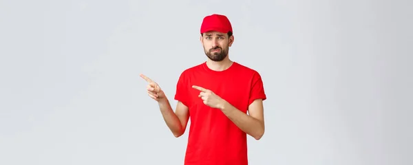 Compras online, entrega durante a quarentena e conceito takeaway. Mensageiro indexicisve e descontente, relutante em t-shirt e boné vermelho, sorrir desapontado, não gostar ideia, apontando os dedos esquerda — Fotografia de Stock