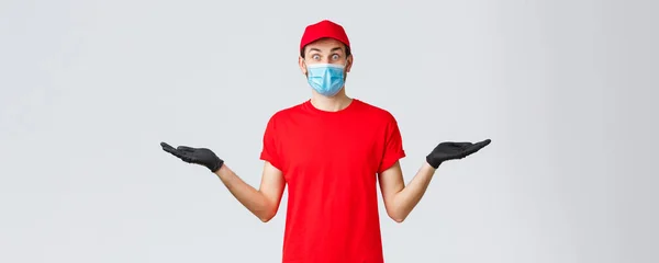 Groceries and packages delivery, covid-19, quarantine and shopping concept. Surprised courier in red uniform, gloves and face mask holding hands sideways with banner or promo of carrier company — Stock Photo, Image