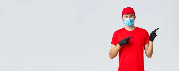 Covid-19, self-quarantine, online shopping and shipping concept. Intrigued smiling delivery guy in red t-shirt, cap carrier uniform, using medical mask and gloves to delivery to client, point right — Stock Photo, Image