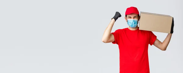 Packages and parcels delivery, covid-19 quarantine delivery, transfer orders. Rejoicing courier in red uniform, face mask and gloves, holding big package box on shoulder and chant, say yes hurray — Stock Photo, Image