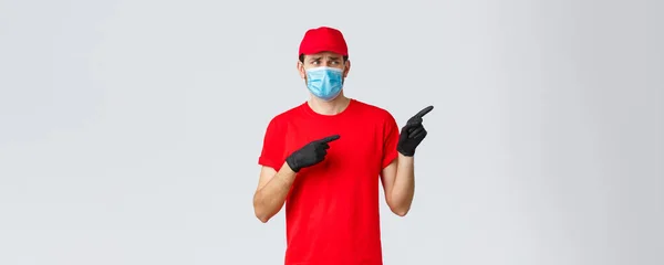 Covid-19, self-quarantine, online shopping and shipping concept. Upset and displeased delivery guy staring and pointing right with reluctant bothered face, wear uniform, medical mask and gloves — Stock Photo, Image