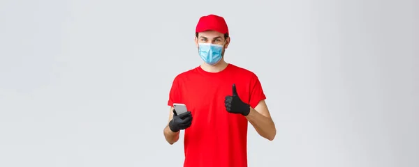 Contactless delivery, payment and online shopping during covid-19, self-quarantine. Friendly courier in gloves and red uniform cap, face mask, show thumb-up as recommend smartphone app or site — Stock Photo, Image
