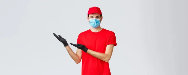 Mercearia e entrega de pacotes, covid-19, quarentena e conceito de compras. homem de entrega amigável em uniforme, máscara facial médica e luvas introduzir nova promo, mostrando bandeira esquerda — Fotografia de Stock