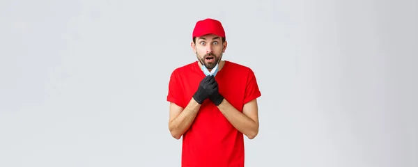 Groceries and packages delivery, covid-19, quarantine and shopping concept. Shocked and speechless courier take-off face mask and drop jaw, staring impressed camera, wear red uniform — Stock Photo, Image