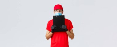Paketler, teslimat, covid-19 karantina, transfer emirleri. Kırmızı üniformalı, eldivenli, maskeli, entrika çeviren, elinde sipariş formu olan meraklı ve heyecanlı kurye.