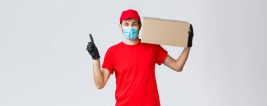 Paketler ve paket teslimatı, karantina teslimatı, nakil emirleri. Yüz maskeli neşeli kurye, eldivenler ve üniforma, paketini kapının önüne taşıyın, yukarı doğru, sırayla kutuyu tutun.