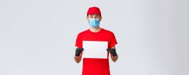 Covid-19, kendini karantina, online alışveriş ve nakliye konsepti. Kırmızı üniformalı, eldivenli ve yüz maskeli, boş kağıtlı heyecanlı bir kurye. Şirket bilgilerini veya afişlerini gösteren şaşkın bir kamera.