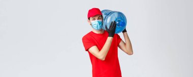 Paket servis, yiyecek ve erzak teslimatı, Covid-19 konsept sipariş konsepti. Kırmızı üniformalı, şapkalı, maskeli ve eldivenli dost canlısı kurye omzunda şişe su siparişi taşıyor.
