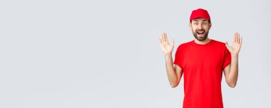 İnternetten alışveriş, karantina sırasında teslimat ve paket servis konsepti. Kırmızı tişörtlü ve şapkalı mutlu kurye, şirket üniforması, eller yukarı şaşkın ve eğlenceli, gri arka plan