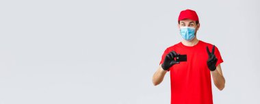 Bağlantı kurulamayan teslimat, ödeme ve kovid 19 sırasında online alışveriş, kendini karantina. Kırmızı üniformalı, eldivenli ve maskeli arkadaş canlısı kurye güvenli ödeme seçeneği sunuyor, kredi kartı ve barış işareti gösteriyor