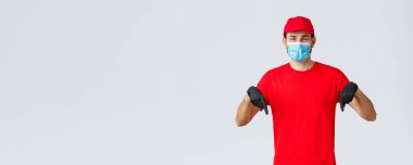 Covid-19, kendini karantina, online alışveriş ve nakliye konsepti. Güler yüzlü teslimatçı adam kırmızı üniformalı, kep ve tişörtlü, eldivenli tıbbi maske takan, yere bakan, tanıtım ve müşteri bilgilerini gösteren