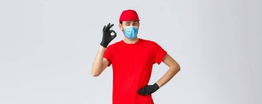 Covid-19, kendini karantina, online alışveriş ve nakliye konsepti. Kırmızı tişörtlü teslimatçı, maske ve eldiven takan şapkalı, tamam işareti göster, göz kırpma, sorun yok de. Herhangi bir transferle kurye ilgilenir