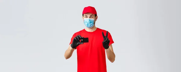 Contactless delivery, payment and online shopping during covid-19, self-quarantine. Friendly courier in red uniform, gloves and face mask provide safe paying option, show credit card and peace sign — Stock Photo, Image
