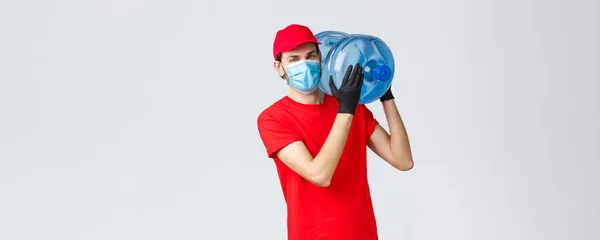 Takeaway, food and groceries delivery, covid-19 contactless orders concept. Young courier in red uniform, cap and face mask with gloves, delivering bottled water to your office or home — Stock Photo, Image