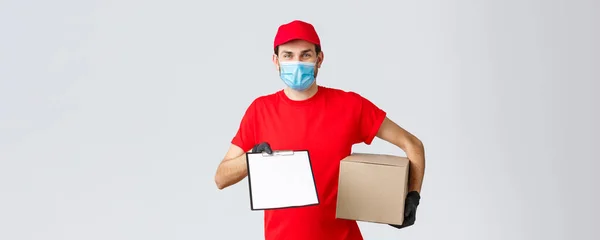 Packages and parcels delivery, covid-19 quarantine delivery, transfer orders. Friendly courier in red uniform, face mask and gloves, holding package box and give clipboard order sign form to client — Stock Photo, Image