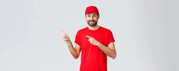 Nätshopping, leverans under karantän- och hämtningskoncept. Vänlig snygg skäggig anställd, kurir i röd uniform mössa och t-shirt, pekfingrar vänster, inbjudande klient ta en titt — Stockfoto