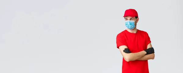 Covid-19, self-quarantine, online shopping and shipping concept. Confident courier in red uniform, protective gloves and face mask, standing crossed arms, look away, ready take your orders — Stock Photo, Image