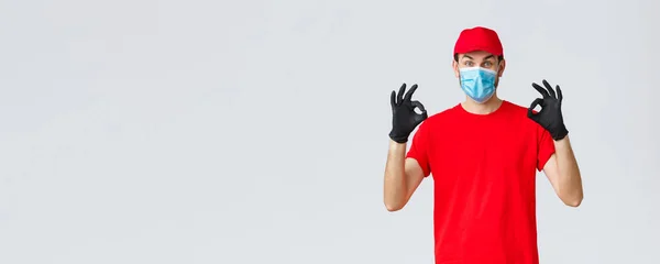 Covid-19, self-quarantine, online shopping and shipping concept. Excited delivery guy in red cap, t-shirt and face mask working coronavirus outbreak, show okay sign, guarantee safe courier service — Stock Photo, Image