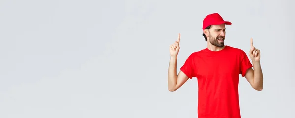 Beau livreur barbu optimiste passer pause détente, employé en uniforme rouge chapeau et t-shirt chanson chantante, pointant les doigts vers le haut insouciant, les yeux fermés, debout fond gris — Photo