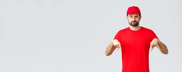 Nätshopping, leverans under karantän- och hämtningskoncept. Skeptisk eller misstänksam kurir i röd uniform mössa och t-shirt, pekar finger ner, stående grå bakgrund — Stockfoto