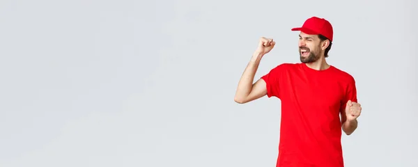 Fröhlicher und unbeschwerter junger bärtiger Lieferant in roter Uniformmütze und T-Shirt, singend, johlend und faustdick in Feierlaune, triumphierend und jubelnd über großartige Neuigkeiten, grauer Hintergrund — Stockfoto