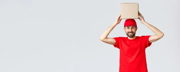 Entrega de pedidos, compras en línea y concepto de envío de paquetes. Divertido y lindo mensajero barbudo en gorra uniforme roja y camiseta, caja de sujeción en la cabeza. Empleado con paquete sonriendo —  Fotos de Stock