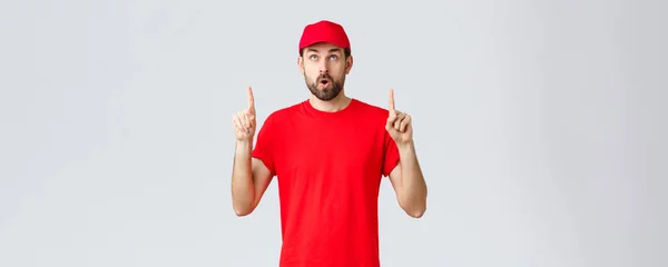 オンラインショッピング、隔離中の配達とテイクアウトの概念。印象的な、赤い制服キャップとTシャツ、口を開けて、興味のあるバナーを読むと、指を指して好奇心の従業員 — ストック写真