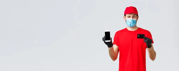 Bezdotykowa dostawa, płatność, zakupy online podczas covid-19, kwarantanna. Przystojny kurier w czerwonym mundurze, rękawiczki i maska, pokazujący ekran smartfona i kartę kredytową, promocja zamówienia — Zdjęcie stockowe