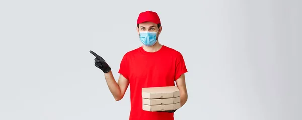 Food delivery, application, online contactless shopping and covid-19 concept. Charismatic courier in red uniform, face mask and gloves, pointing finger left, holding pizza, bring order client — Stock Photo, Image