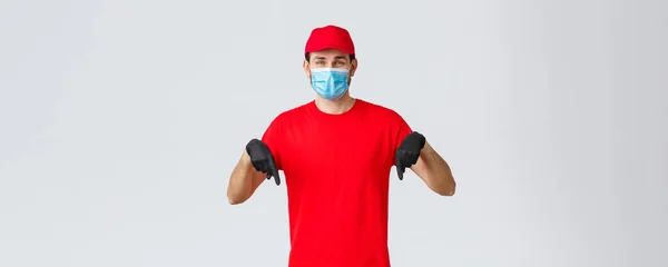 Covid-19, self-quarantine, online shopping and shipping concept. Smiling friendly delivery guy red uniform, cap and t-shirt, wear medical mask with gloves, pointing down, showing promo or client info — Stockfoto