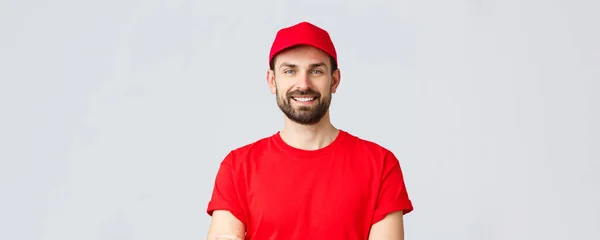 Online shopping, delivery during quarantine and takeaway concept. Handsome bearded courier in red uniform, smiling cheerful, cross arms chest, ready to deliver your order, grey background — Stock Photo, Image