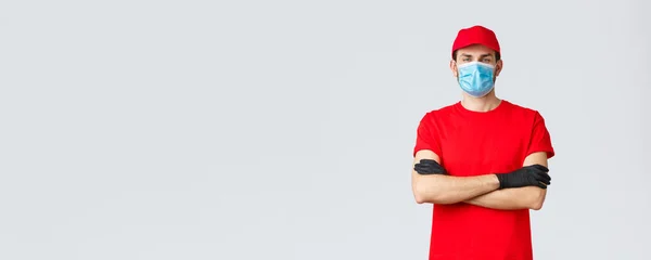 Pewny siebie młody dostawca w czerwonej koszulce i czapce, noszący maskę medyczną i rękawice ochronne podczas dostarczania zamówień klientom przebywającym w domu bezpiecznie podczas kwarantanny dla koronawirusów — Zdjęcie stockowe