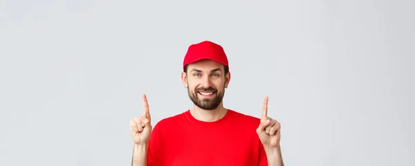 オンラインショッピング、隔離中の配達とテイクアウトの概念。陽気な髭の笑顔の宅配便で赤の制服キャップとTシャツ,プロモーションを見てみましょう,指を指して,灰色の背景 — ストック写真