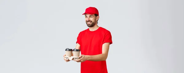 Livsmedelsleverans, karantän, stanna hemma och beställ online koncept. Vänlig kurir i röd uniform mössa och t-shirt, dela ut kaffe till kunder, ta med drinkar via drive-through fönster, grå bakgrund — Stockfoto