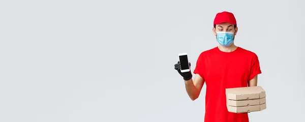Food delivery, application, online grocery, contactless shopping and covid-19 concept. Excited courier in red uniform, looking at pizza boxes amused, showing smartphone screen app or bonus promo — Stock Photo, Image