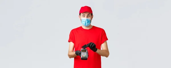 Contactloze levering, betaling en online winkelen tijdens covid-19, self-quarantaine. Spannende koerier in rood uniform, gezichtsmasker en handschoenen, kijk verrast, met behulp van een creditcard op het betalen van terminal POS — Stockfoto