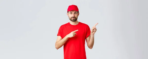 オンラインショッピング、隔離中の配達とテイクアウトの概念。赤い制服キャップとTシャツに自信を持って髭を生やした宅配便,笑顔のカメラ,方向を与える,指の右を指して,クライアントに通知 — ストック写真