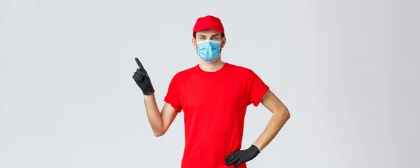 Covid-19, self-quarantena, shopping online e concetto di spedizione. Uomo di consegna in berretto rosso e t-shirt, maschera medica con guanti per proteggere clienti e dipendenti, puntare il dito sinistro al promo, mostrare annuncio — Foto Stock