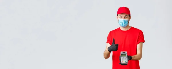 Contactless delivery, payment and online shopping during covid-19, self-quarantine. Friendly smiling courier in red uniform cap, t-shrit, medical mask and gloves, advice pay order with POS terminal — Stock Photo, Image