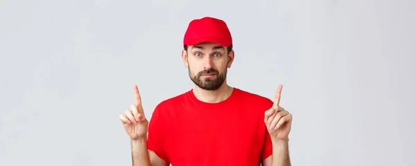 オンラインショッピング、隔離中の配達とテイクアウトの概念。赤の制服キャップ、 Tシャツ、笑顔と指を指差す不確定で不確定な宅配便、わからない、できない決定 — ストック写真
