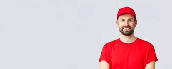 Compras en línea, entrega durante la cuarentena y concepto de comida para llevar. Guapo mensajero barbudo en uniforme rojo, sonriente alegre, pecho de brazos cruzados, listo para entregar su pedido, fondo gris — Foto de Stock